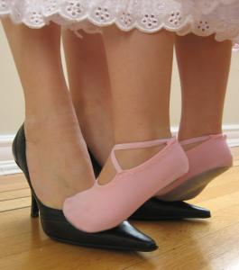 A little girl in pink ballet slippers stands on her mother's feet.