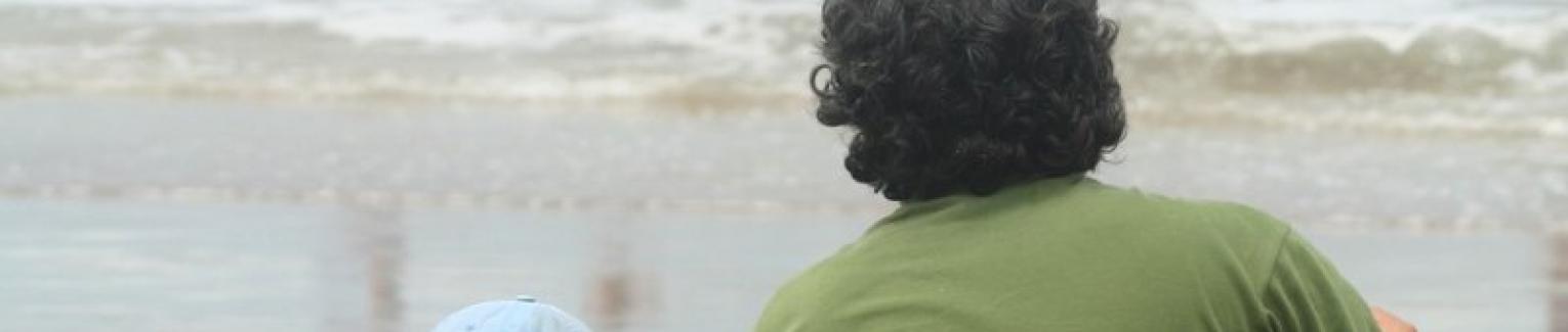 A Father sitting on the beach with his small child, watching the ocean surf.